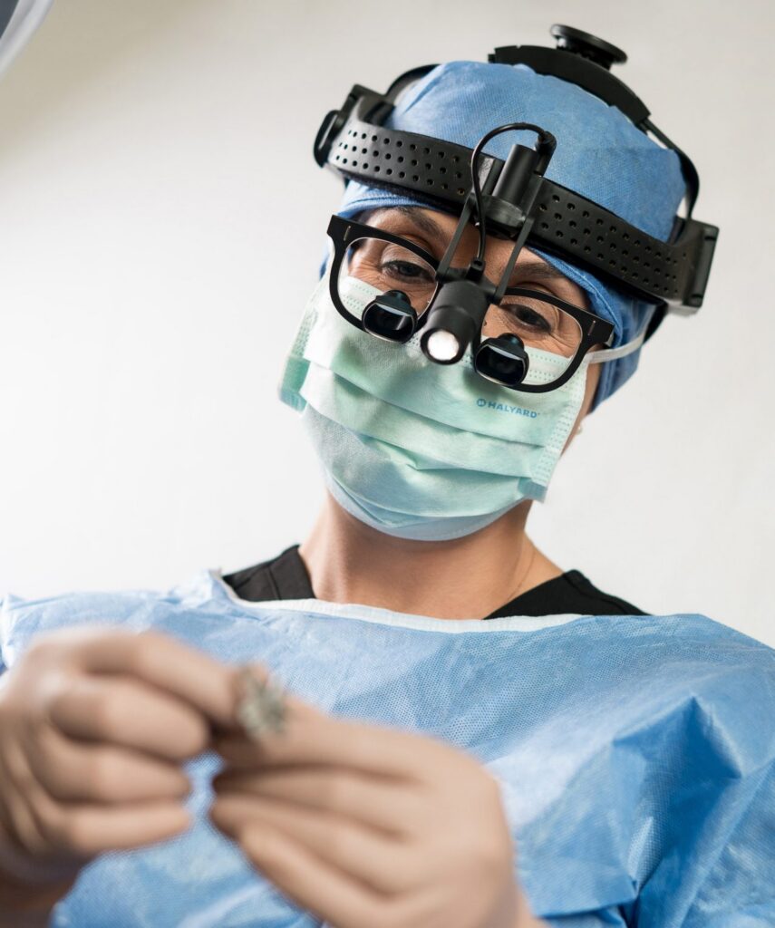 Dr. Caitlin Magraw in surgical loupes and a mask, examining a small surgical implant